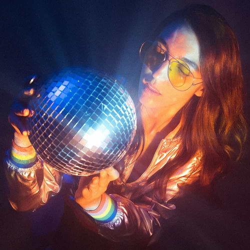 A woman wearing yellow-tinted sunglasses and a shiny metallic jacket holds a glowing disco ball, surrounded by colorful, moody lighting.