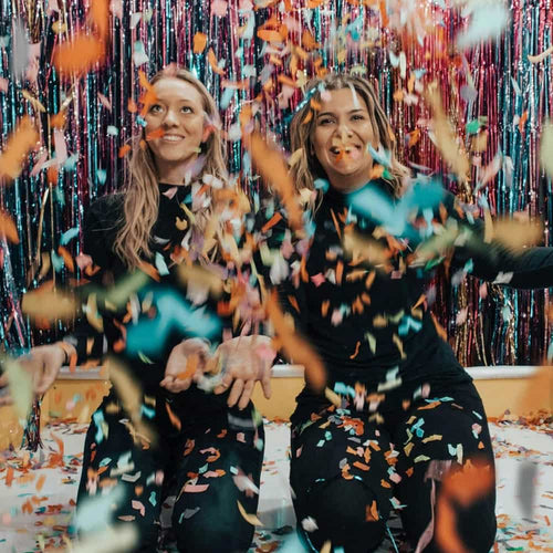 Two women in black outfits sit in front of shiny, colorful streamers as they toss vibrant confetti into the air, smiling and celebrating together.