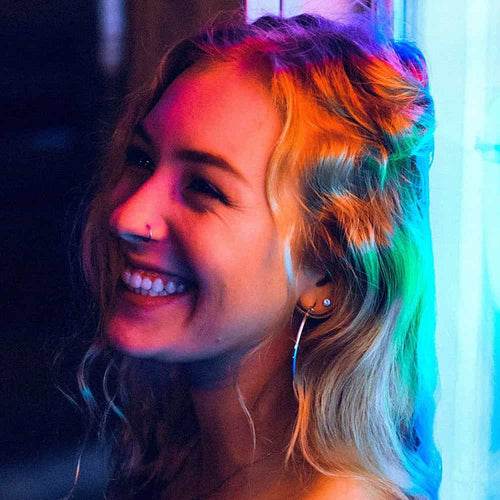 A smiling young woman with wavy blonde hair stands near neon lights, creating colorful reflections on her face. She has a nose ring and hoop earrings, and is looking to the side, appearing joyful.