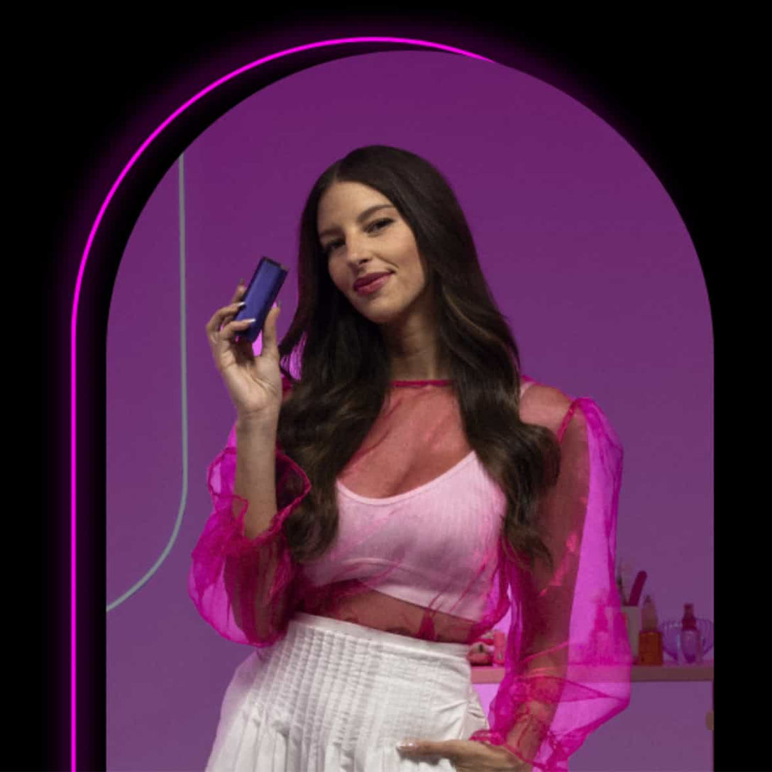 A woman with long dark hair, wearing a sheer pink top over a white undershirt and a white skirt, stands confidently holding a small rectangular device, set against a purple background framed by a glowing pink arch.