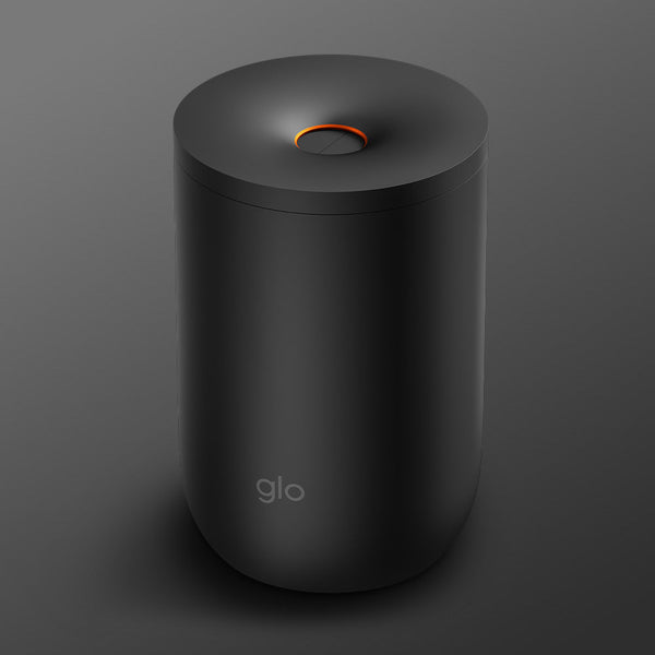 The glo™ Stick Bin is a sleek, cylindrical matte black device with a subtle glo logo near the bottom, a circular orange accent on top, and comes with a premium fabric cover for extra protection, shown against a gray background.