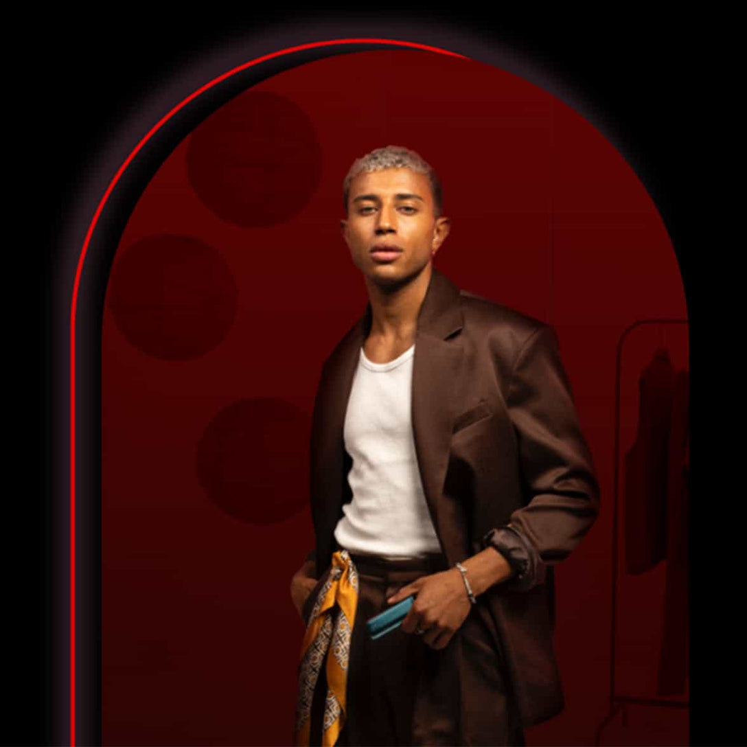 A stylish person with short, light hair stands in a dark archway, wearing a brown suit jacket over a white shirt, brown pants, and a yellow patterned scarf tied at the waist, holding a small blue item.