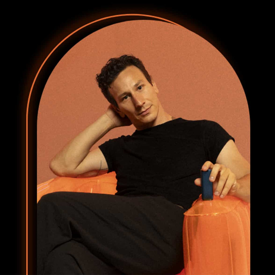 A man in a black shirt and pants sits on an orange inflatable chair, resting his head on one hand and holding a small rectangular object in the other, against a peach-colored background.