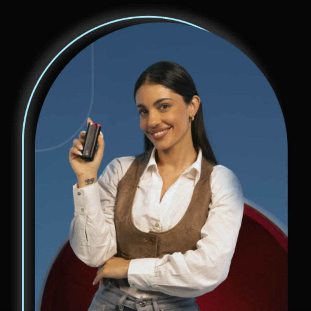 A woman with long dark hair, wearing a white shirt and brown vest, smiles while holding up a small black device in her right hand against a blue background with a red curved chair.