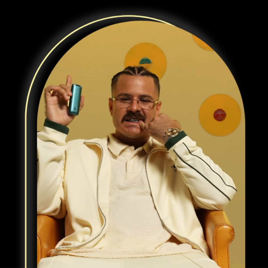 A man in a cream tracksuit with dark stripes sits in a tan chair against a yellow background with orange circles, holding a teal device in one hand and touching his face with the other. He has glasses, a mustache, and slicked-back hair.